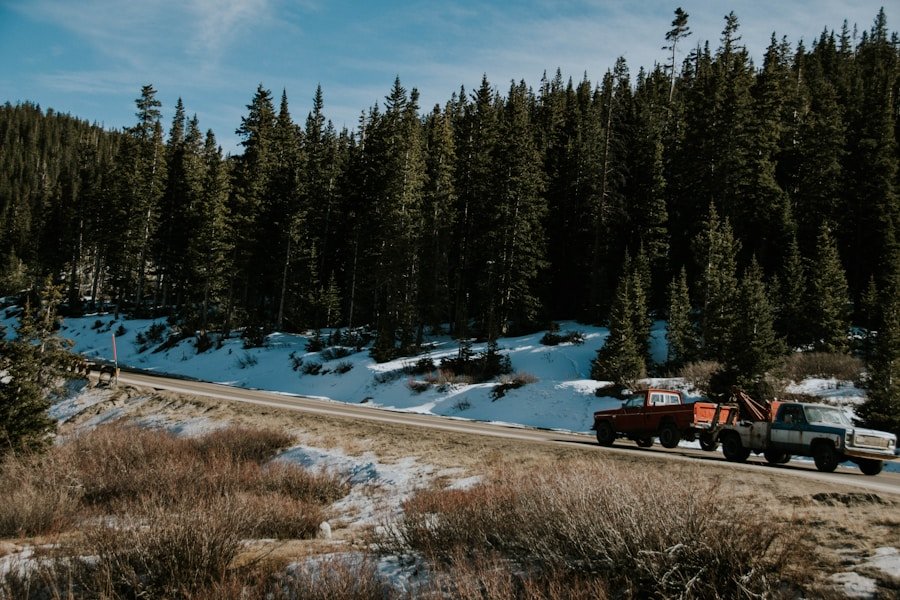 A tow truck pulls another truck on a snow-dusted road beside a forest of evergreen trees.